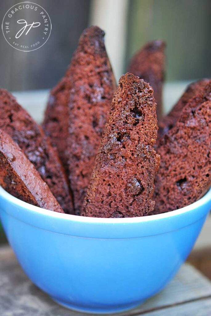 A side view of a blue bowl filled with chocolate biscotti sit on a wooden table.