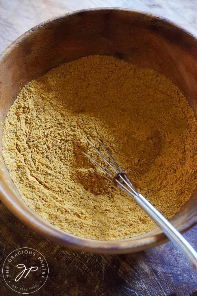 Jamaican Curry Powder sits in a wooden bowl with a whisk resting on the side of the bowl.