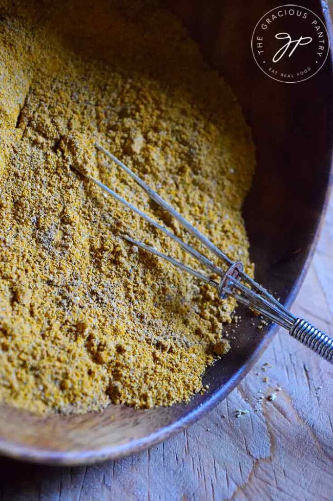 A wooden bowl sits tipped on its side. A small whisk rests in the bowl, sitting half on the counter and half in the Jamaican Curry Powder in the bowl.