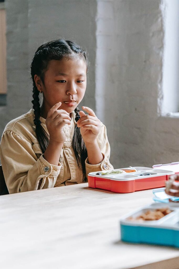 A young girl sits at a lunch table, eating with her lunchbox in front of her.