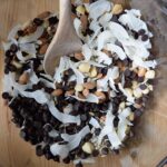 A clean mixing bowl with a wooden spoon resting in it. All the ingredients for this Low Carb Trail Mix Recipe sit in the bowl for mixing.