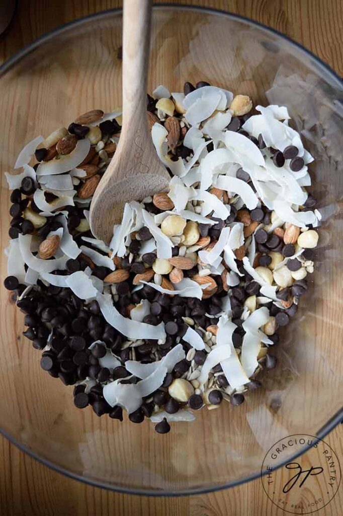 A clean mixing bowl with a wooden spoon resting in it. All the ingredients for this Low Carb Trail Mix Recipe sit in the bowl for mixing.