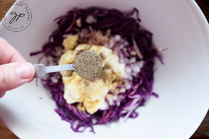 Holding a measuring spoon of black pepper over the mixing bowl to add to this red cabbage slaw recipe.