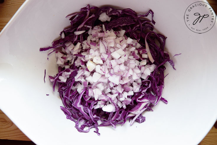 The chopped red onions added to the cabbage in a white mixing bowl.
