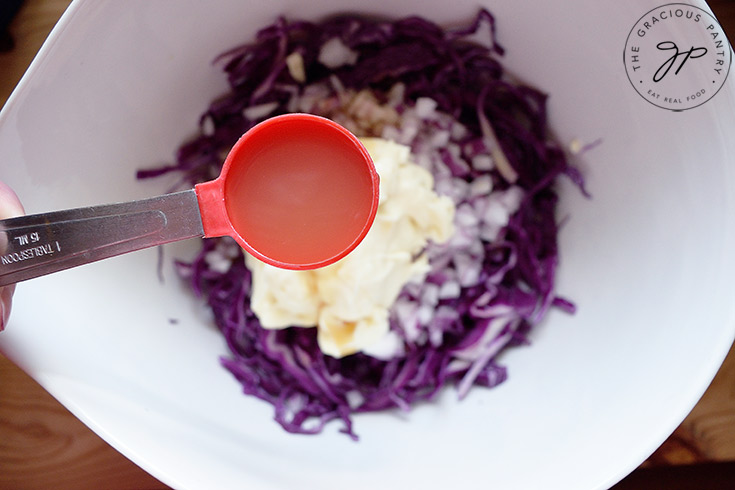 Holding a tablespoon measure of apple cider vinegar over the mixing bowl to add to this Red Cabbage Slaw Recipe.