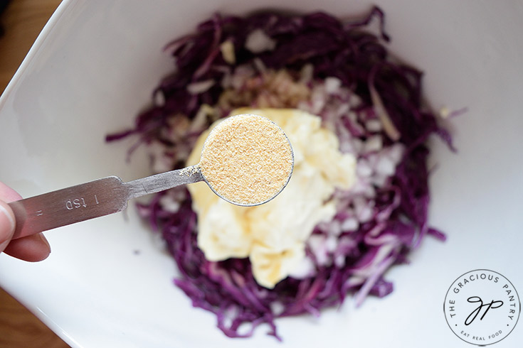 Holding a teaspoon measure of garlic powder over the mixing bowl to add to this Red Cabbage Slaw Recipe.