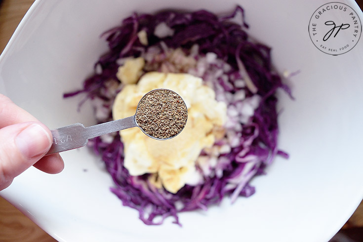 Holding a half teaspoon measure of ground black pepper over the mixing bowl to add to this Red Cabbage Slaw Recipe.