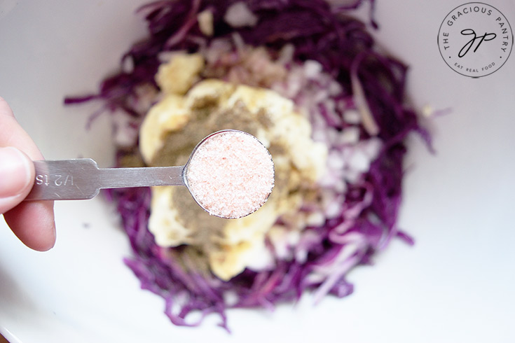 Holding a half teaspoon measure of salt over a white mixing bowl to add to this Red Cabbage Slaw Recipe.