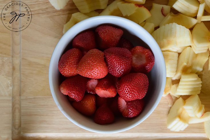 A bowl of fresh strawberries sits on a cutting board, surrounded by chopped apples.