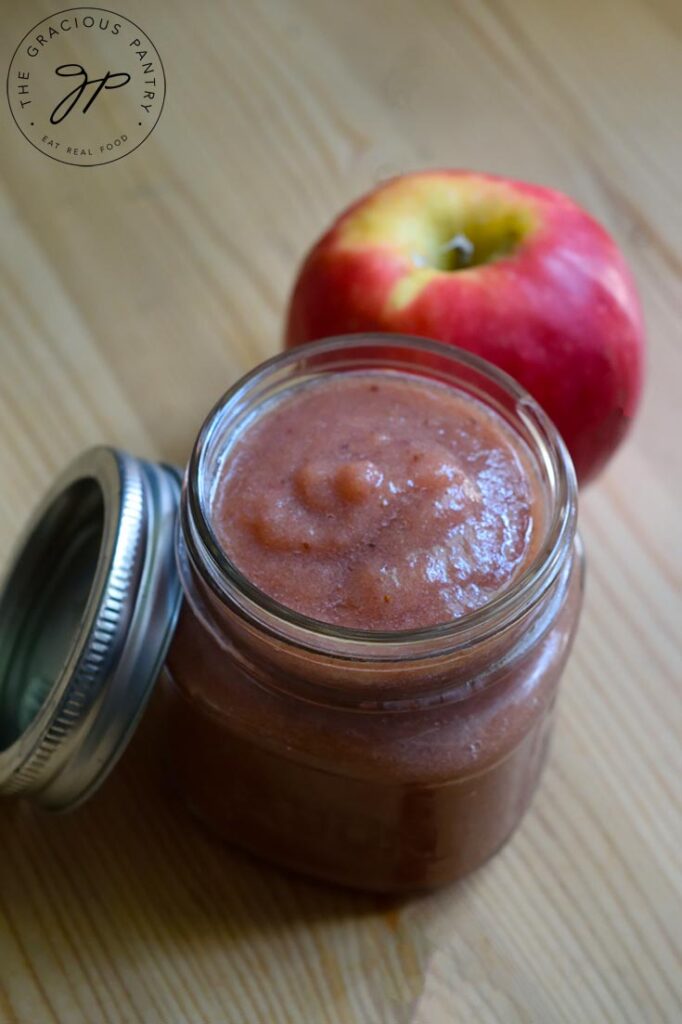 An overhead shot of a canning jar filled with Strawberry Apple Sauce. The jar lid and a whole apple sit to the side of the jar.