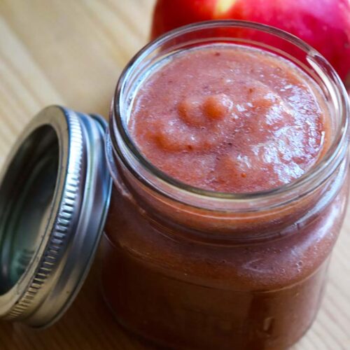 An open canning jar filled with Strawberry Apple Sauce sit with a lid to the left and an whole apple behind the jar.