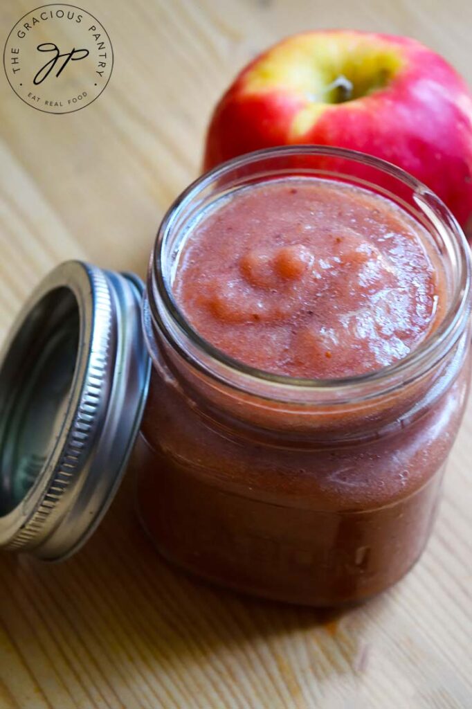 An open canning jar filled with Strawberry Apple Sauce sit with a lid to the left and an whole apple behind the jar.