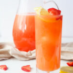 A tall, skinny glass, filled with Strawberry Lemonade and garnished with lemon and strawberry slices.