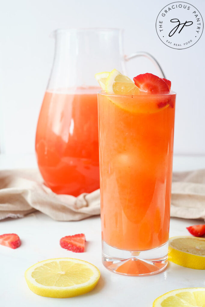 A tall, skinny glass, filled with Strawberry Lemonade and garnished with lemon and strawberry slices.