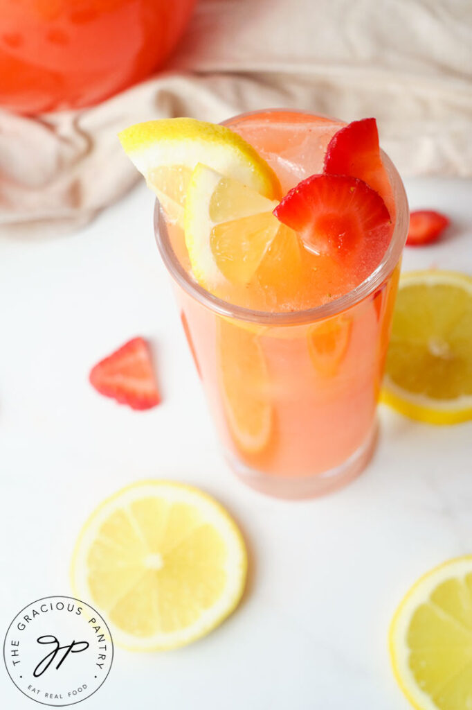 A overhead shot looking down into a tall glass filled with this Strawberry Lemonade Recipe. A pitcher stands behind it and a few lemon slices are scattered around the glass on the table.