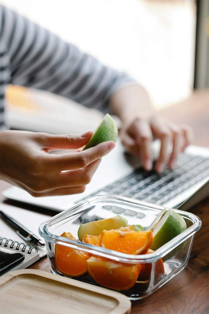 Person at a computer eating fresh apple and orange slices in this guide to Healthy Snacks For Teachers