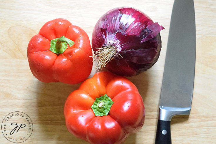 A red onion and two red bell peppers sit next to a knife on a wooden cutting board.