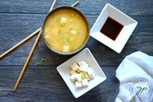 Componants of this Miso Soup Recipe in separate bowls. Tamari, cubed tofu, and a finished bowl of soup on a gray background. Chopsticks lay to the side.
