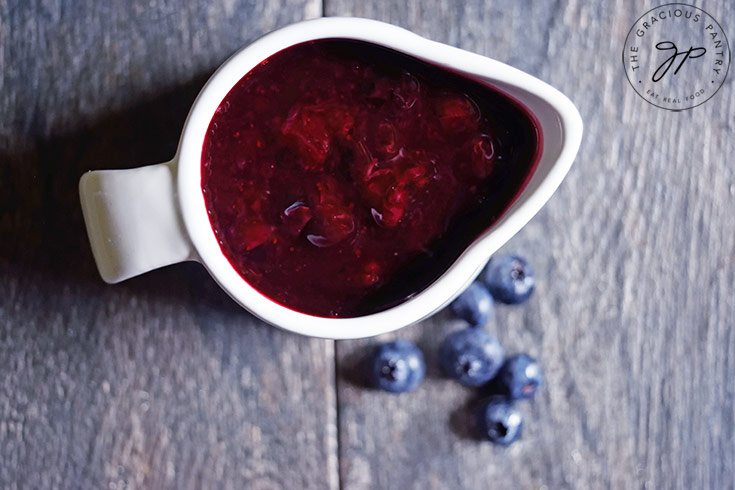 The finished blueberry syrup in a white pitcher. A few fresh blueberries lay to the side of the pitcher.