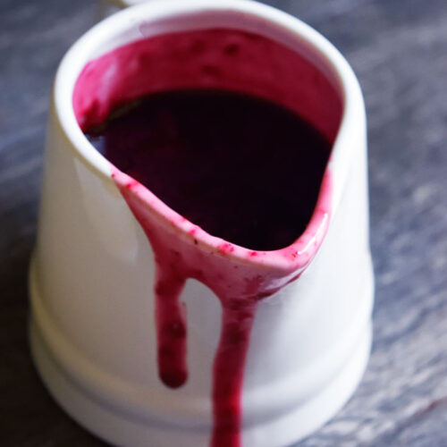 A single white pitcher filled with Blueberry Syrup. A bit of syrup has spilled over the edge of the pitcher and is dripping down the front.