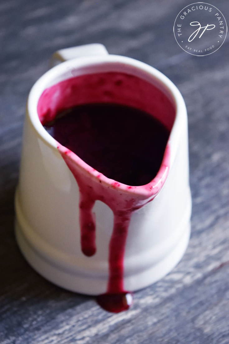 A single white pitcher filled with Blueberry Syrup. A bit of syrup has spilled over the edge of the pitcher and is dripping down the front.