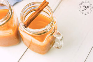 An overhead view looking down into two mugs of Pumpkin Cider. A cinnamon stick rests over the top of the mugs.