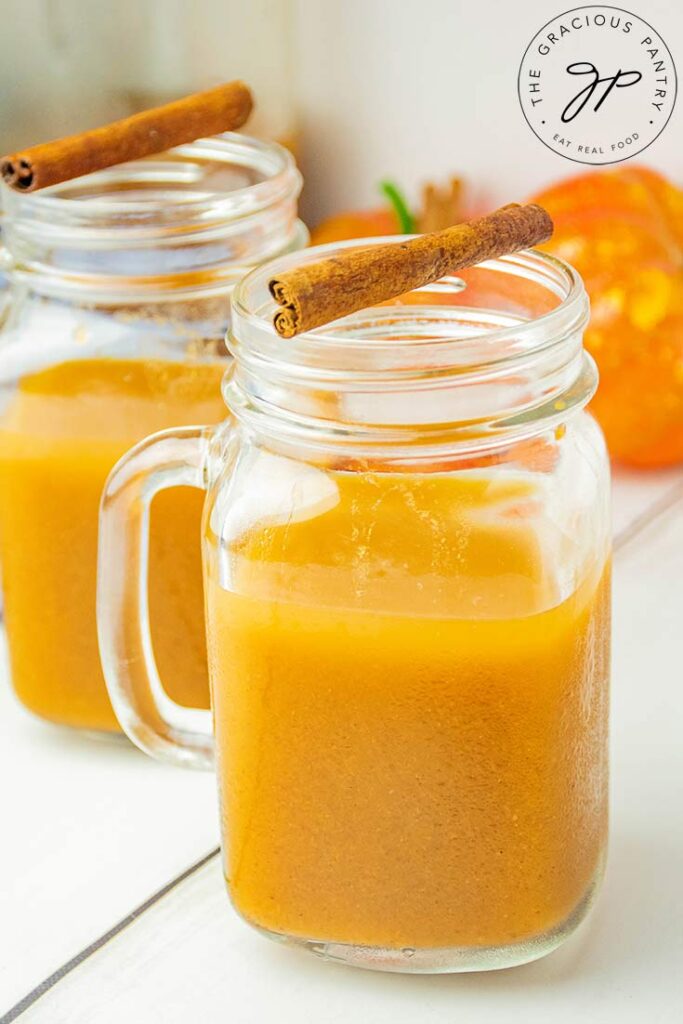 Two mugs of pumpkin cider. A cinnamon stick rests on the top of both mugs.