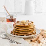 A plate of Pumpkin Oatmeal Pancakes sits on a white table with white jars and glasses sitting behind it.