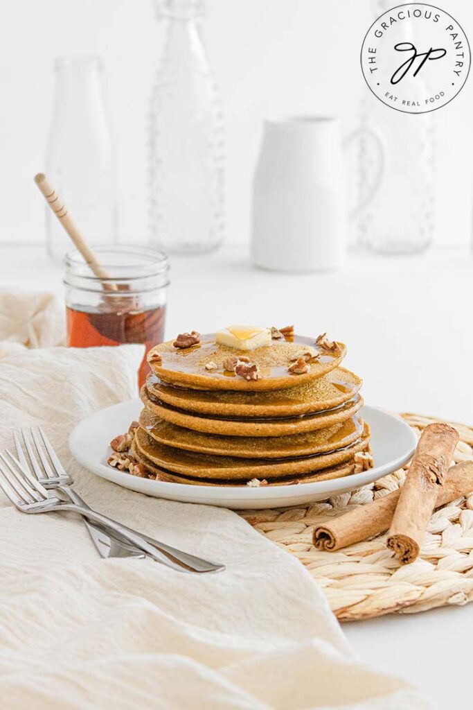 A plate of Pumpkin Oatmeal Pancakes sits on a white table with white jars and glasses sitting behind it.