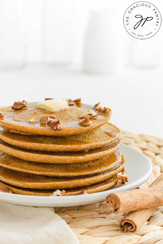 And up close side shot of a stack of Pumpkin Oatmeal Pancakes on a white plate. Two cinnamon sticks sit to the front right of the plate.