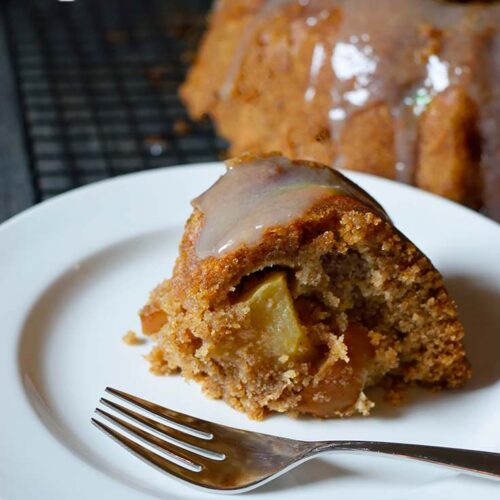 A slice of Spiced Apple Bundt Cake sits on a white plate. A fork rests to the side of the slice.