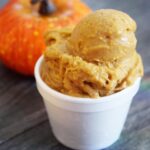 A scoop of Pumpkin Ice Cream in a white bowl with a small pumpkin sitting behind the cup.