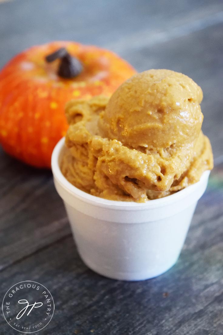 A scoop of Pumpkin Ice Cream in a white bowl with a small pumpkin sitting behind the cup.