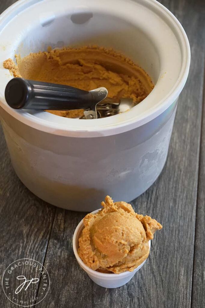 An ice cream scoop sits in the ice cream maker insert. In front of it, sits a small white bowl of just-scooped Pumpkin Ice Cream.