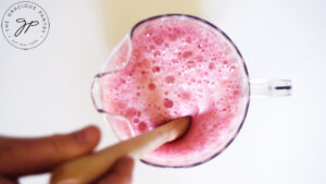 A full pitcher with all the ingredients for this Cranberry Mimosa Mocktail Recipe being stirred by a wooden spoon.