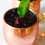 Two copper mugs filled with this Pomegranate Moscow Mule Mocktail Recipe and garnished with a fresh cranberry and mint leaf.