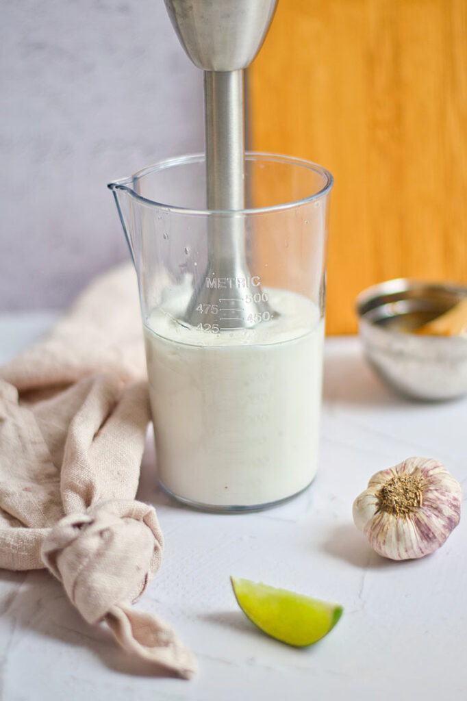 An immersion blender sits in a clear tumbler filled with just blended aioli sauce.