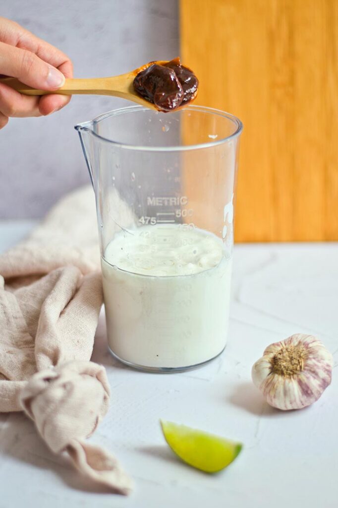 A measure of chipotle peppers being added to a clear tumbler with aioli in it.