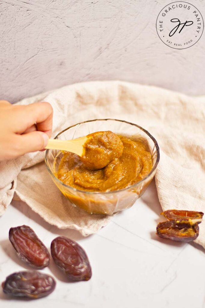 A woman's hand scooping some date caramel out of a glass bowl with a small, wooden spoon.