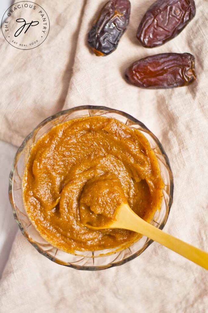 And up close view from overhead of a bowl filled with date caramel. A small wooden spoon rests in the caramel.