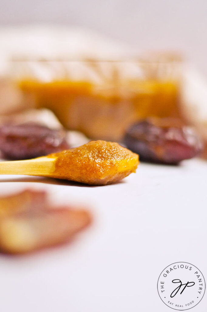 A side view of a small wooden spoon with date caramel on it, resting on a white table top.