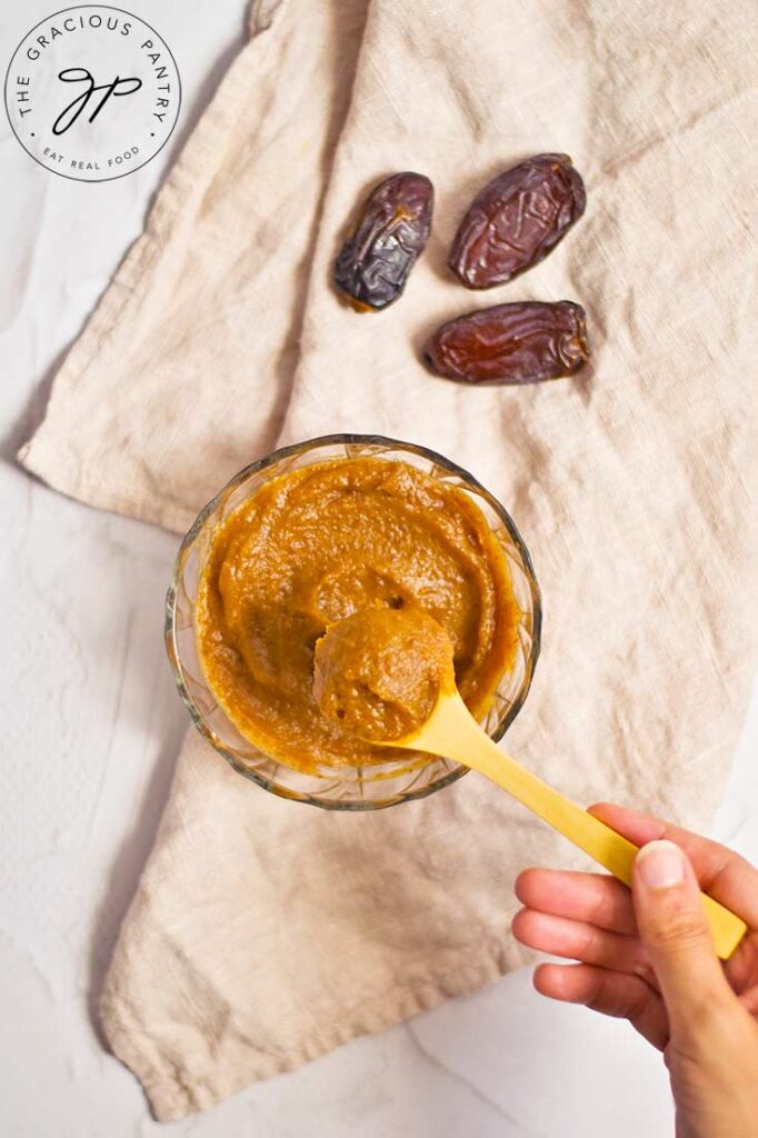 And overhead view of a female hand lifting a spoonful of date caramel out of a serving bowl and towards the camera.