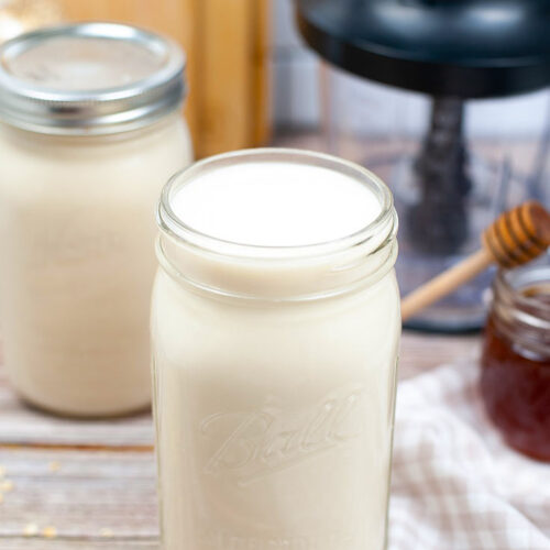 A canning jar filled with oat milk sits with it's top off, next to a blender and two other closed jars of oat milk.