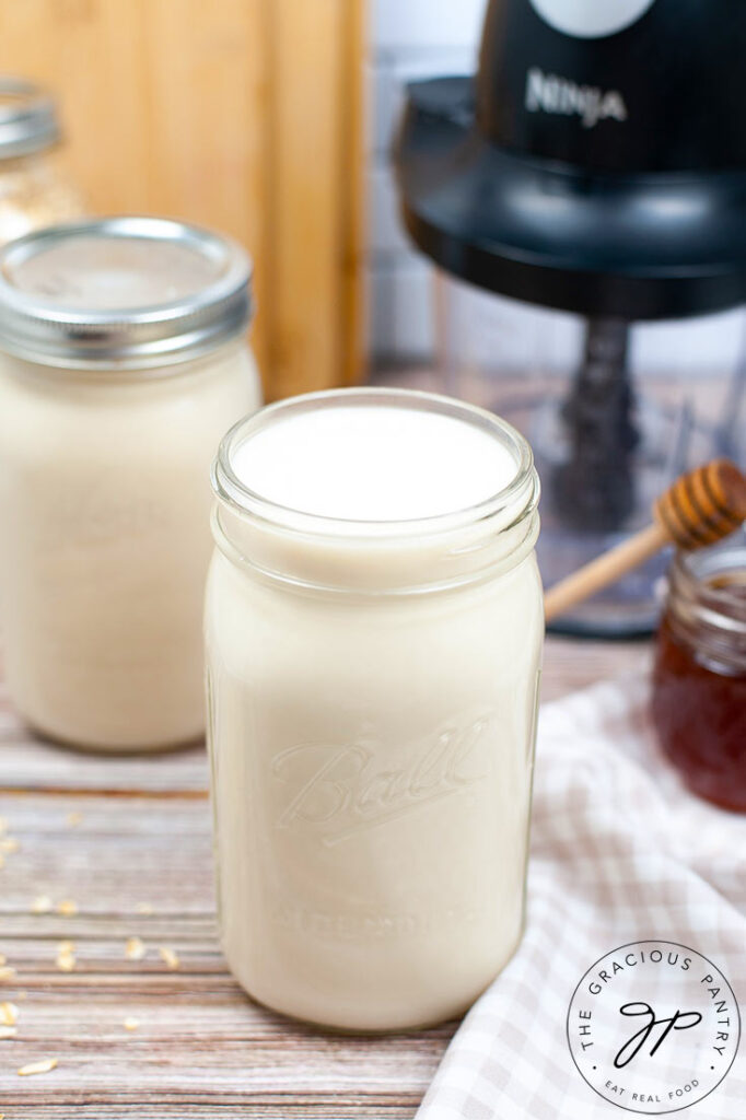 A canning jar filled with oat milk sits with it's top off, next to a blender and two other closed jars of oat milk.