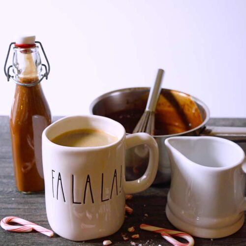 A gathering of dishes. A mug of coffee, a small white pitcher, a pot and a jar filled with this Protein Peppermint Mocha Creamer Recipe. Broken candy canes lay on the table in front of the mug.