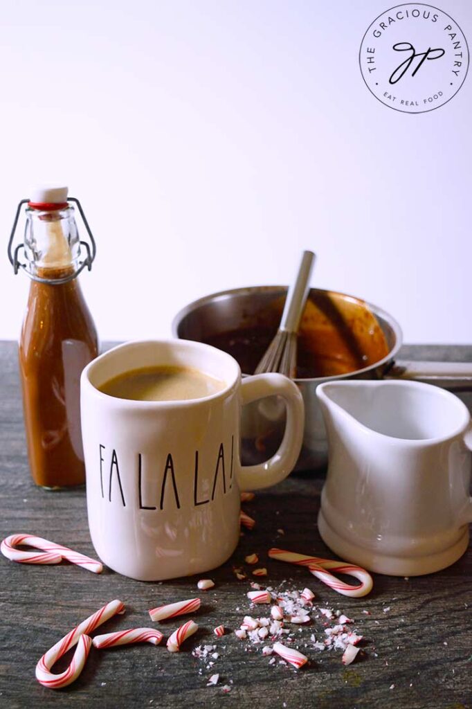 A gathering of dishes. A mug of coffee, a small white pitcher, a pot and a jar filled with this Protein Peppermint Mocha Creamer Recipe. Broken candy canes lay on the table in front of the mug.