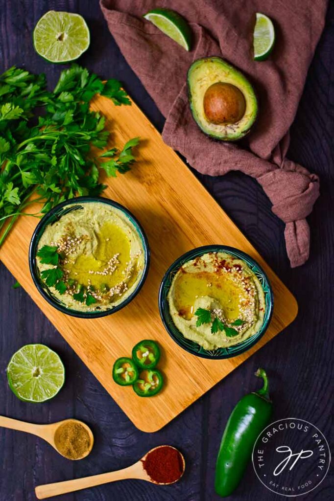 A dark table holds a cutting board which has two black bowls filled with Avocado Hummus. Various ingredients lay around the cutting board.