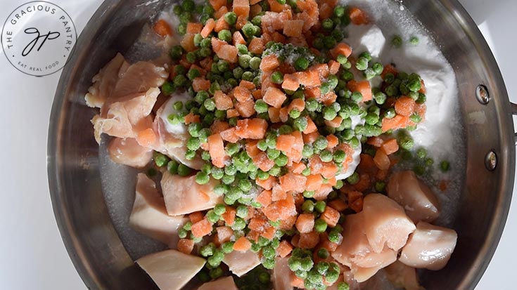 Frozen peas and carrots top chicken and coconut milk in a skillet.