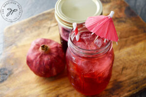 The finished Grenadine Syrup in a canning jar, a shirley temple drink and a whole pomegranate sit on a cutting board.