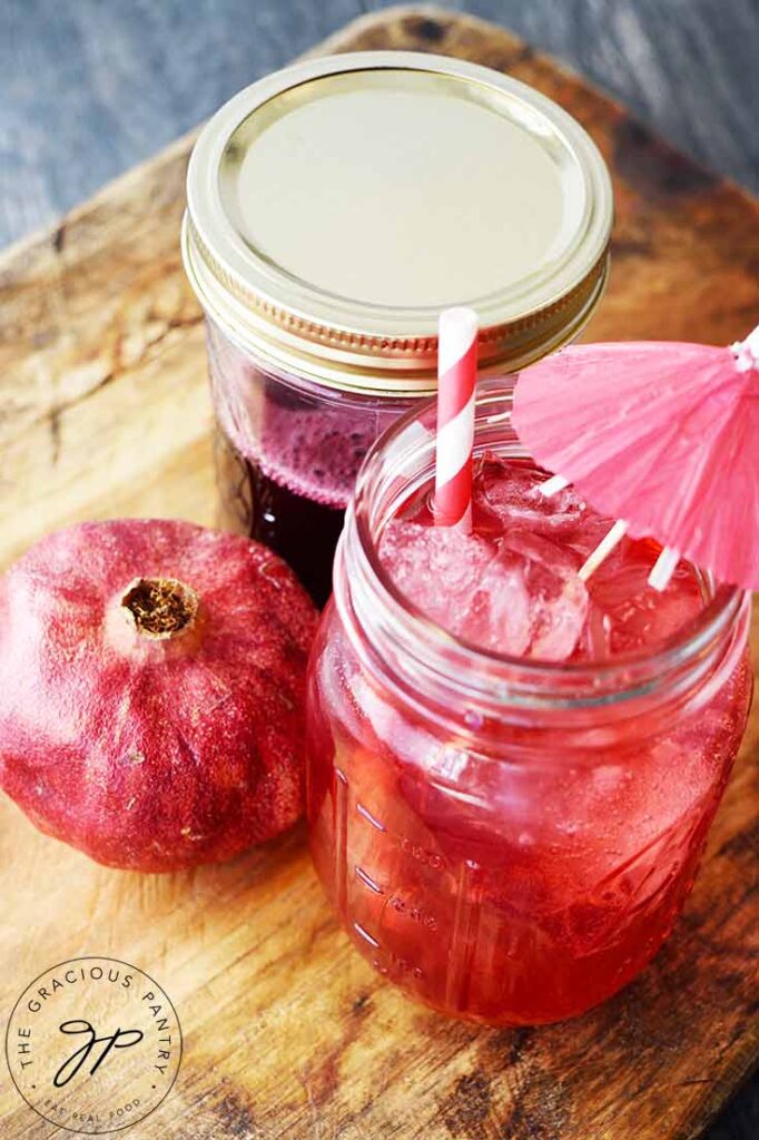 An overhead view looking down on a cutting board that holds a glass of shirley temple, a whole pomegranate ad a jar of Grenadine Syrup.